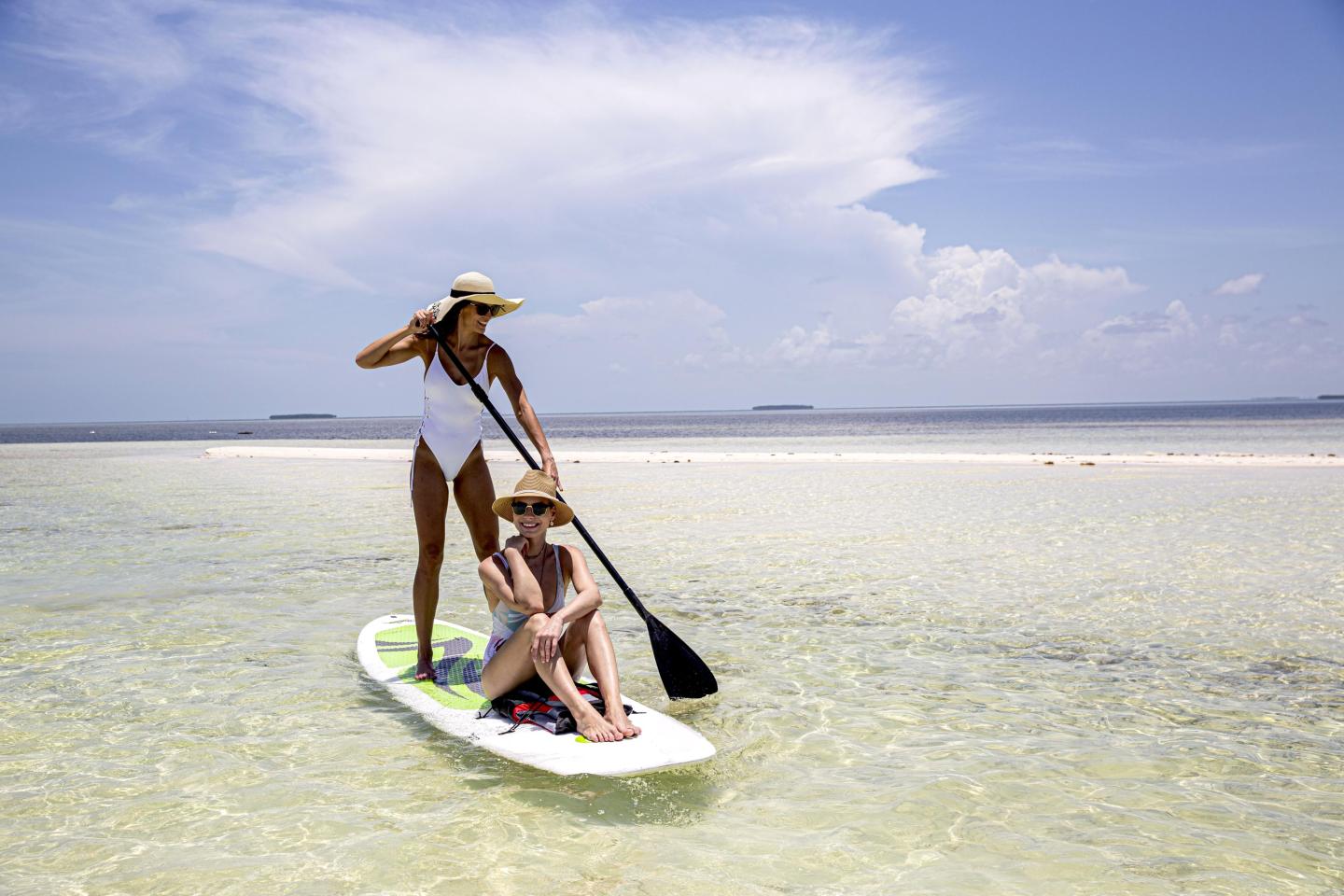 Two women paddleboarding on clear, shallow water under a blue sky.
