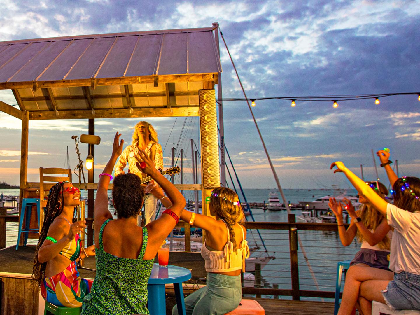 Outdoor gathering by the sea at sunset, people dancing and cheering near a stage.