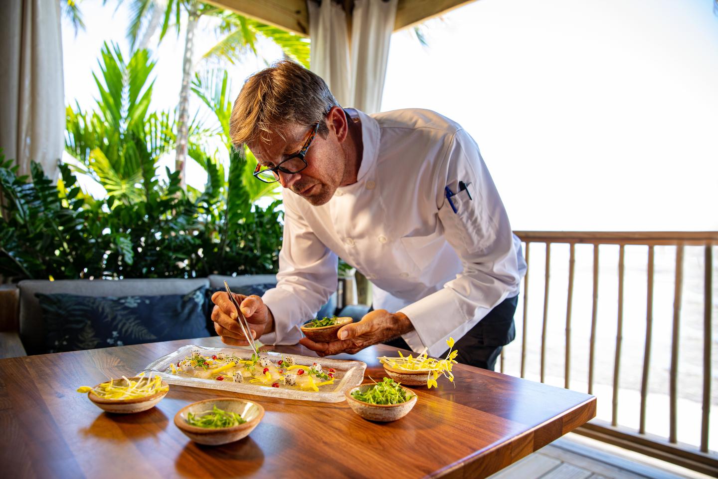 Chef plating food on a wooden table outdoors, surrounded by greenery.