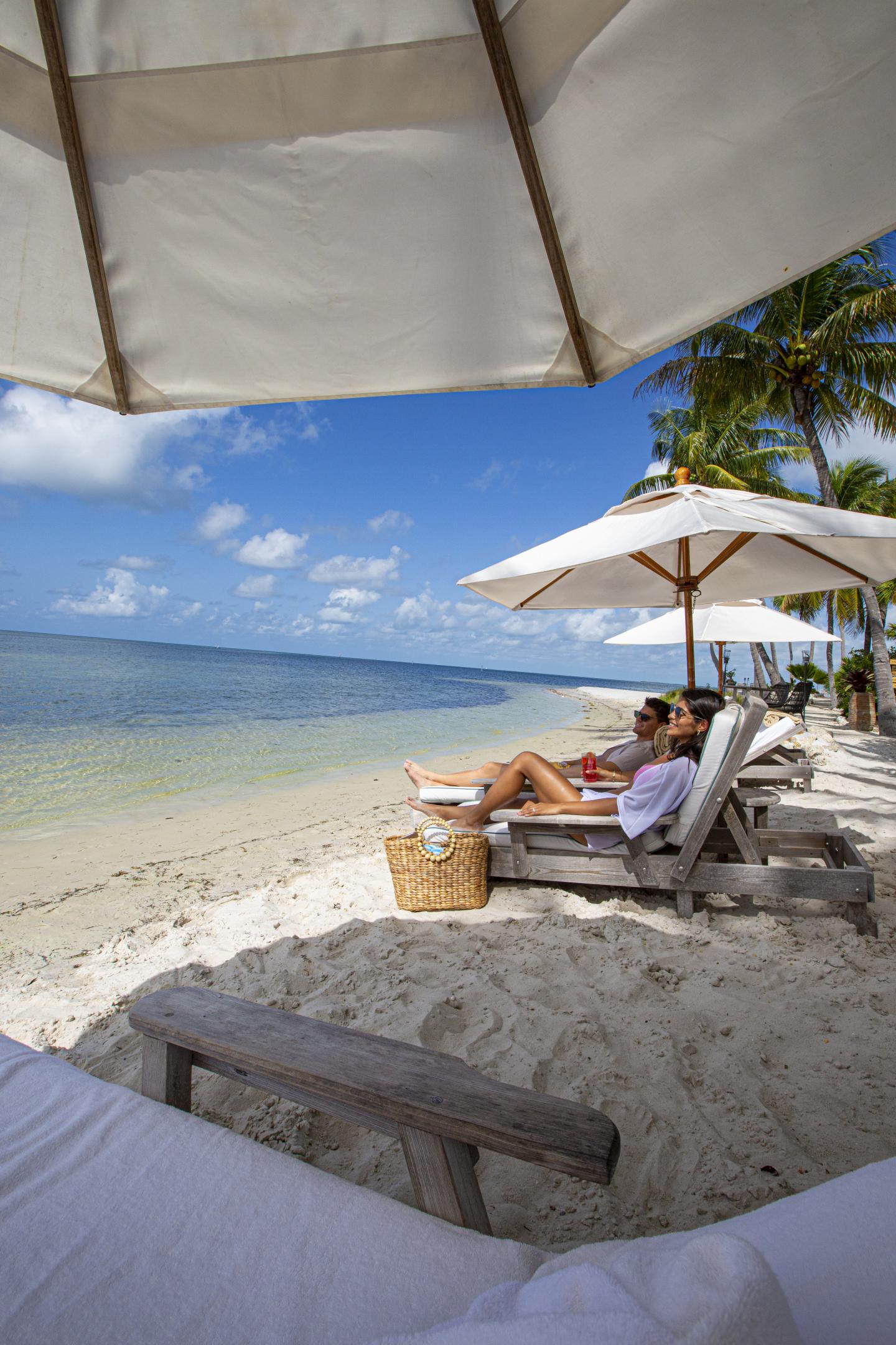 Beach scene with lounge chairs, umbrellas, and palm trees by the ocean.