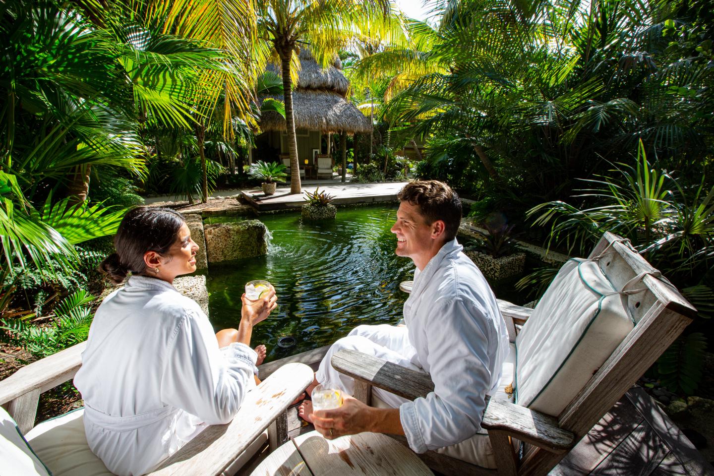 Two people in bathrobes relax by a tropical pond, surrounded by lush greenery.