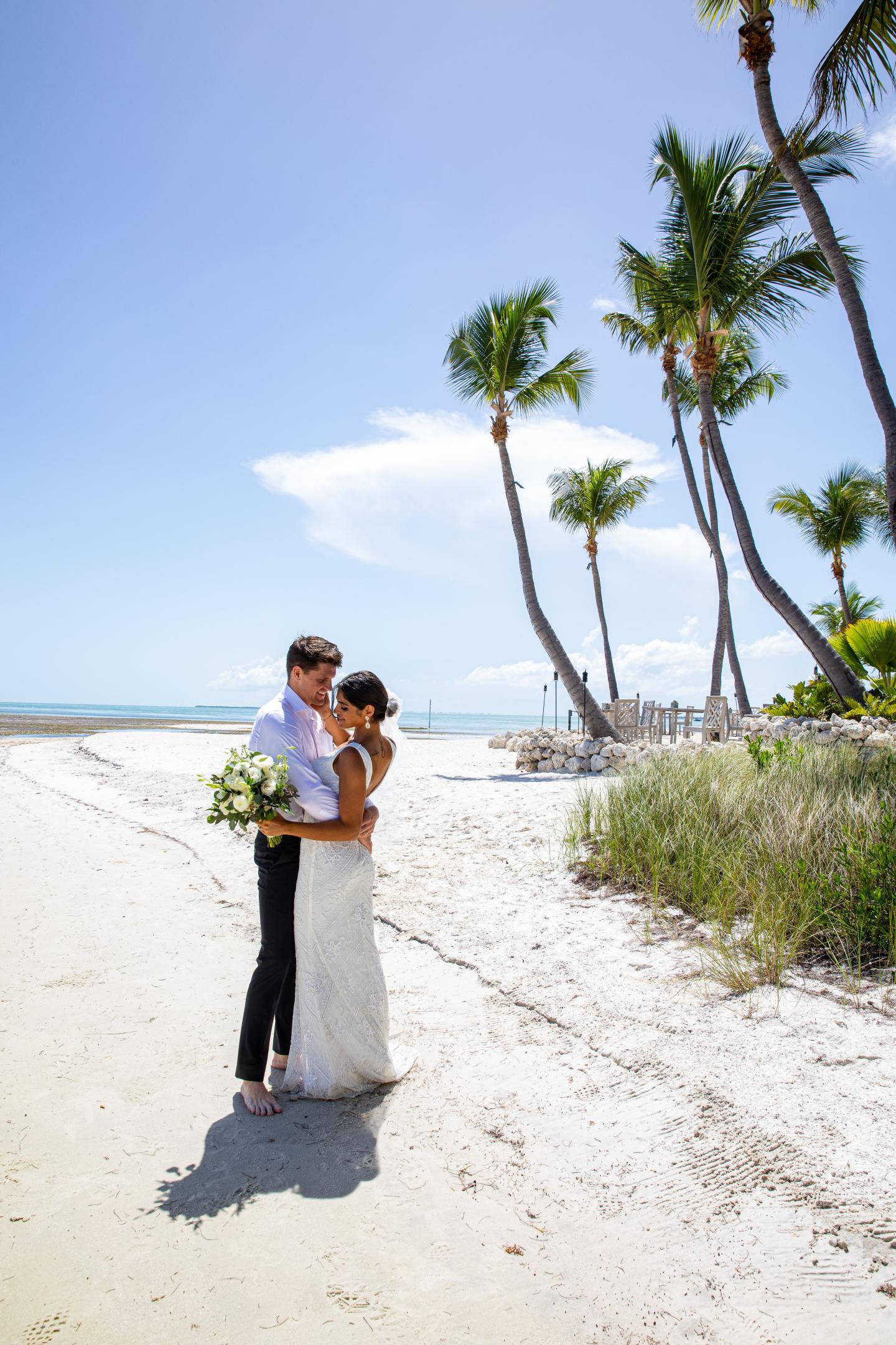 Bride and groom embrace on a sunny beach with palm trees.