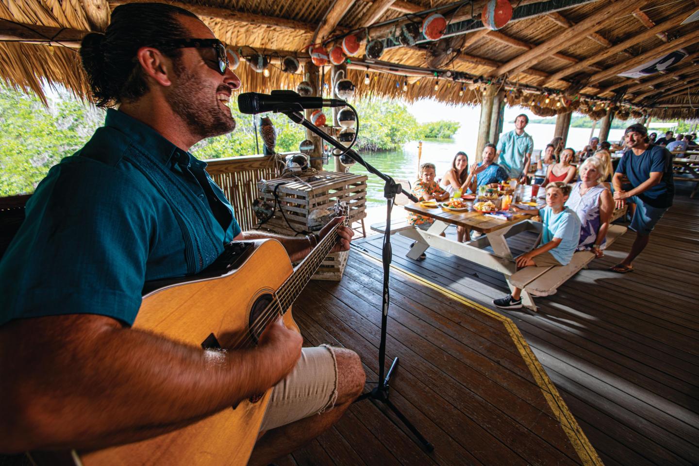 Musician playing guitar, singing at a sunny outdoor restaurant, people dining nearby.