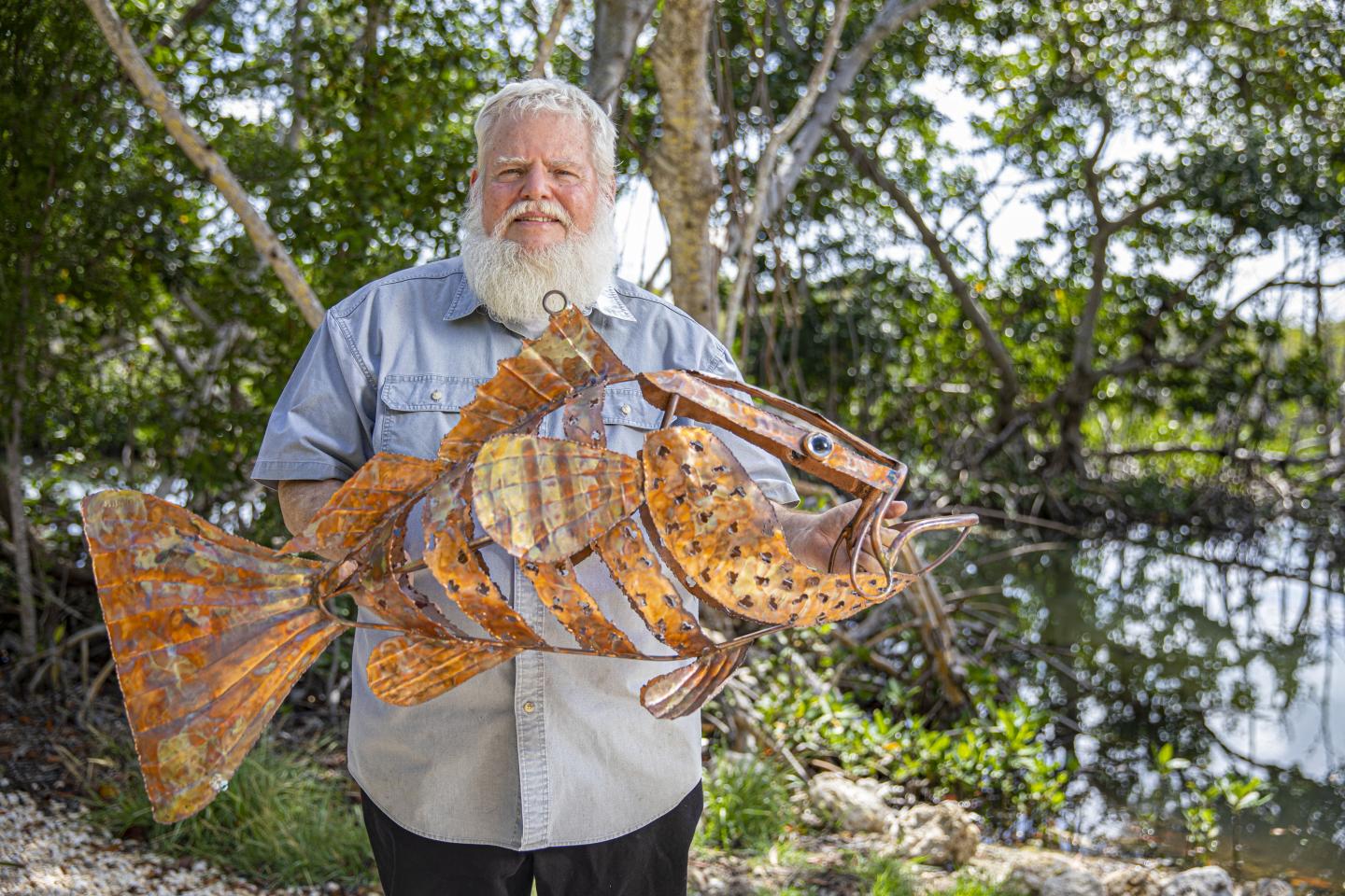 Bearded man holding a large, rust-colored metal fish sculpture outdoors.