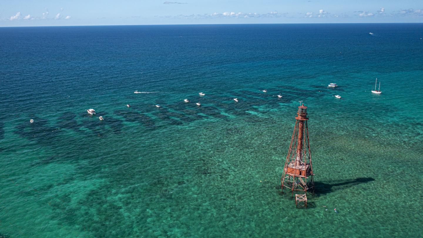 Lighthouse on clear turquoise sea with boats in the distance.