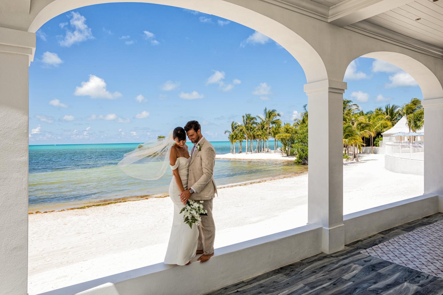 Bride and groom kiss by a sunny beach under an arched balcony.