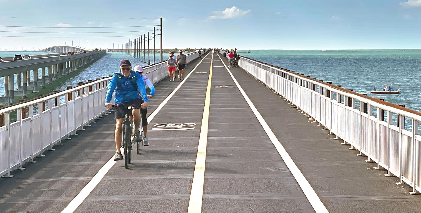 Cyclists riding on a long bridge over the sea, under a clear blue sky.