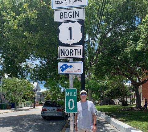 A man stands by a road sign for Mile 0, Route 1 North, under a tree.