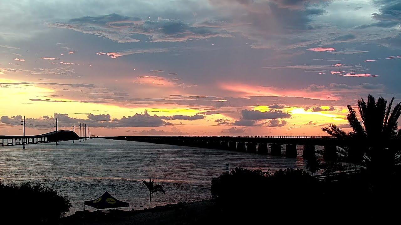 Sunset over a bridge and water, with silhouettes of trees and a boat.