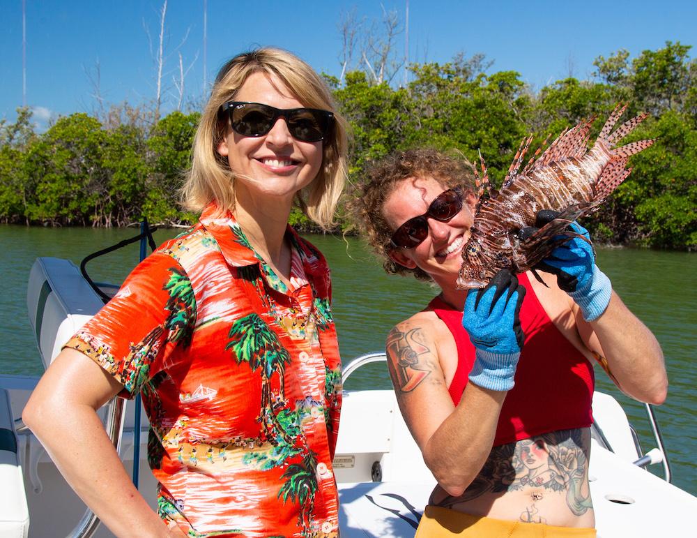 Two people smiling on a boat; one holds a lionfish.
