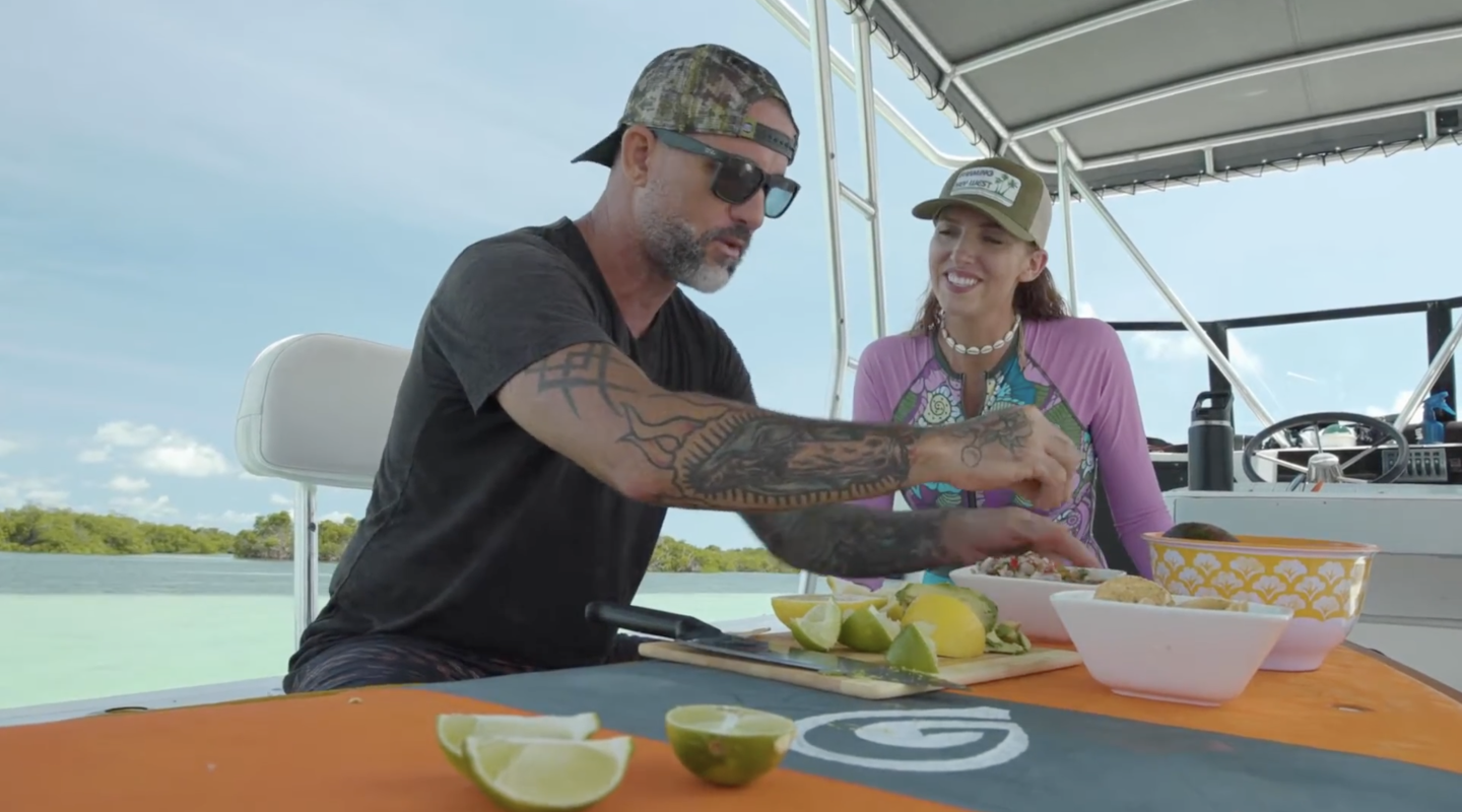 Man slicing lime on a boat while woman watches, both wearing caps.