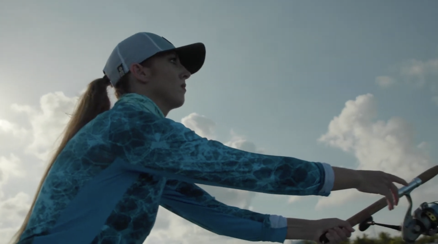 A person in a blue patterned shirt and cap fishing under a cloudy sky.