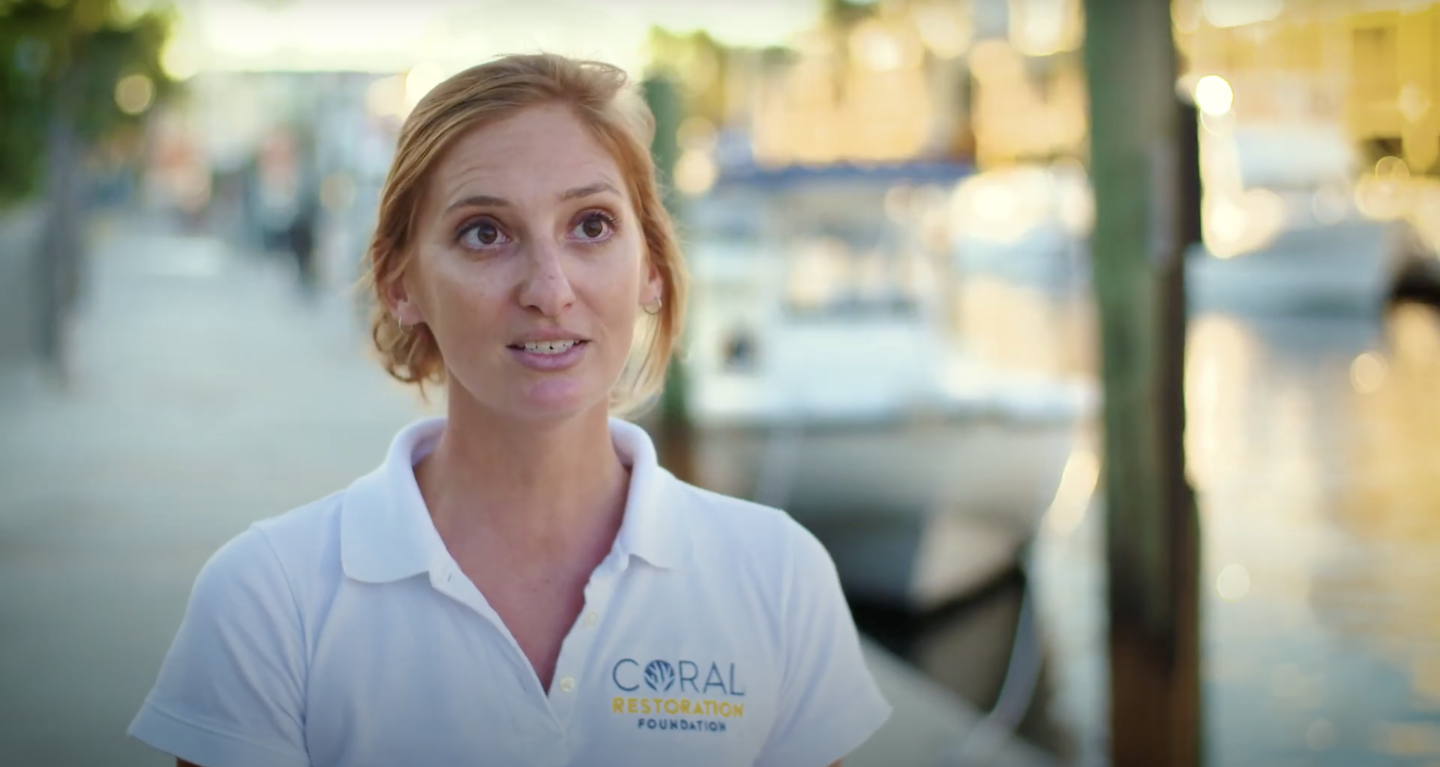 Woman smiling by a marina, wearing a white shirt. Blurred boats in the background.