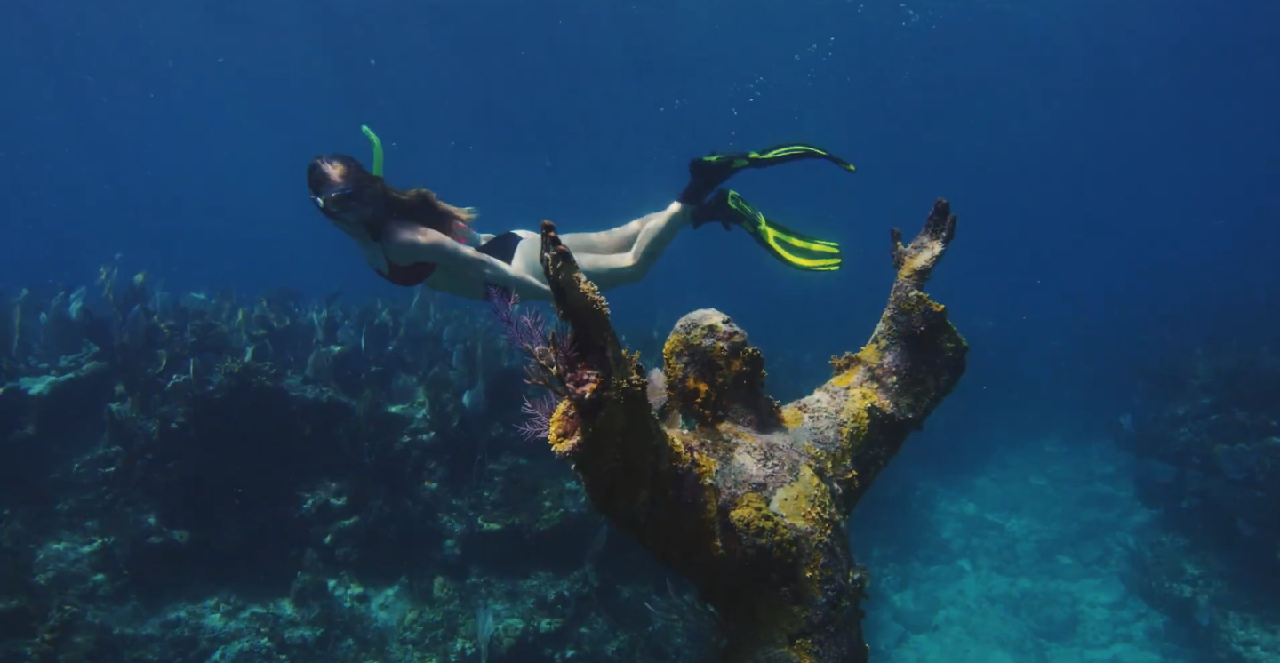 Underwater statue with diver swimming nearby, vibrant corals, deep blue sea.