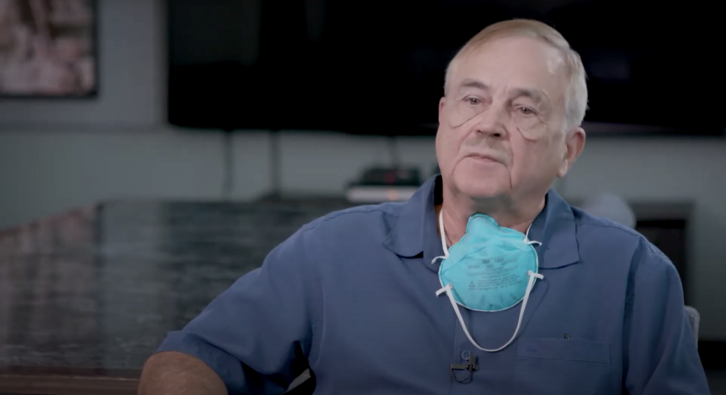 Middle-aged man wearing a blue shirt and teal mask around his neck indoors.