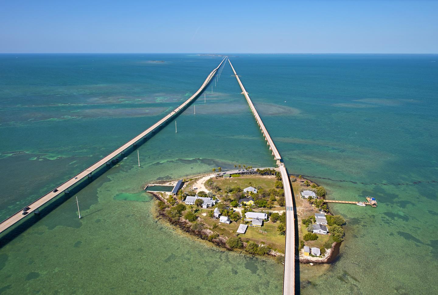 Aerial view of a long bridge over turquoise ocean, with a small island below.