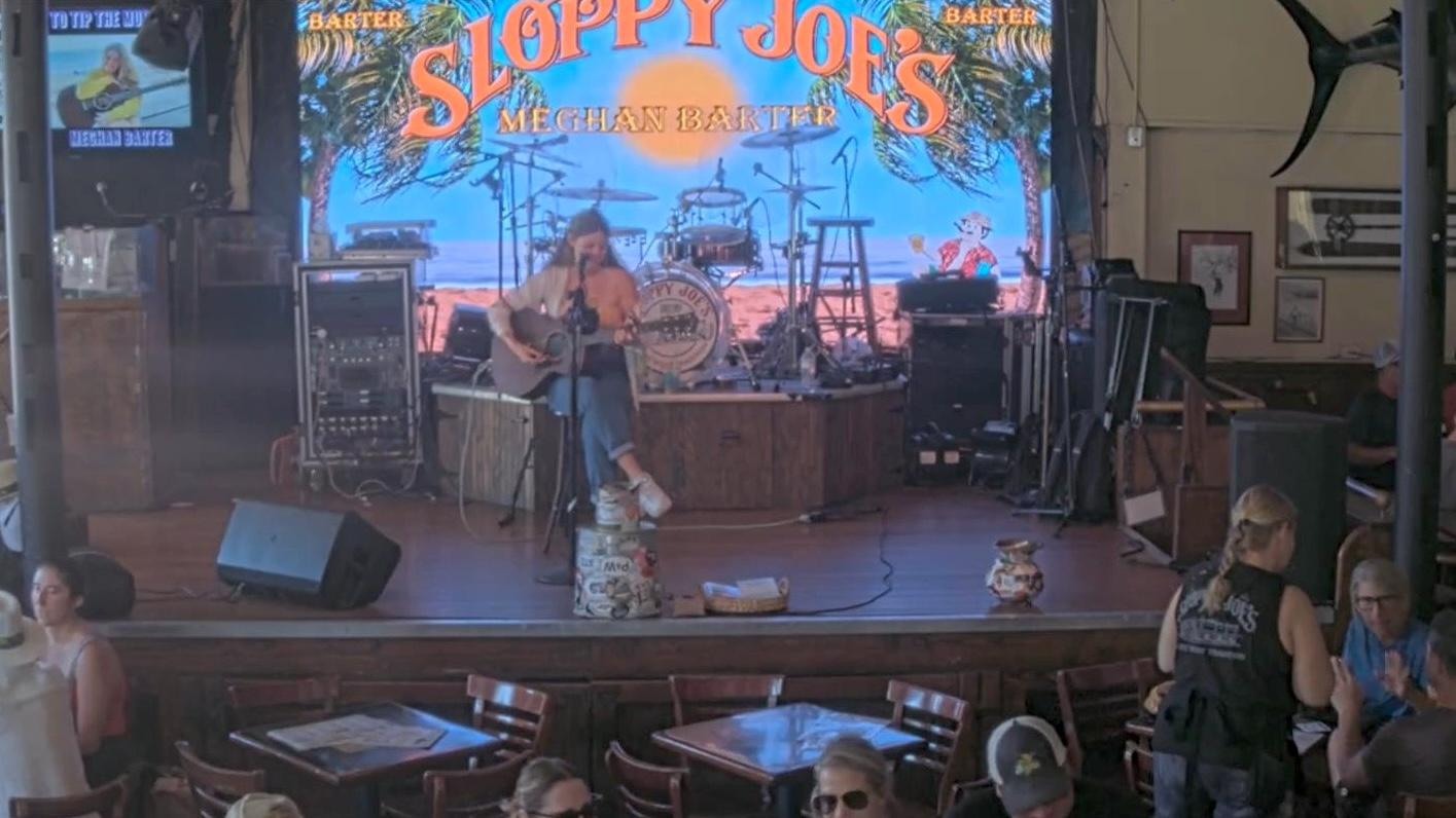 Musician performing on stage at Sloppy Joe's, people seated at tables.