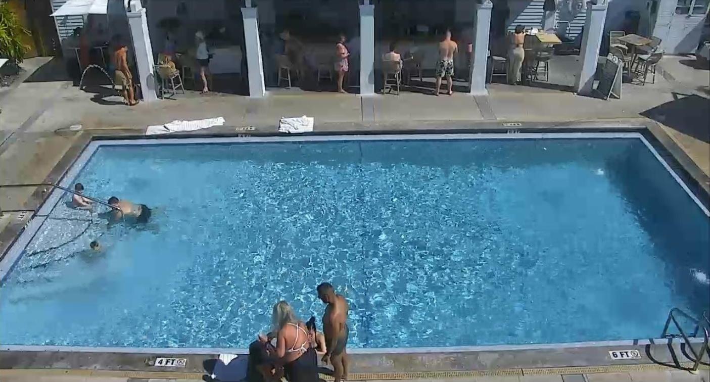 Outdoor pool with people swimming and sunbathing; bar in background.