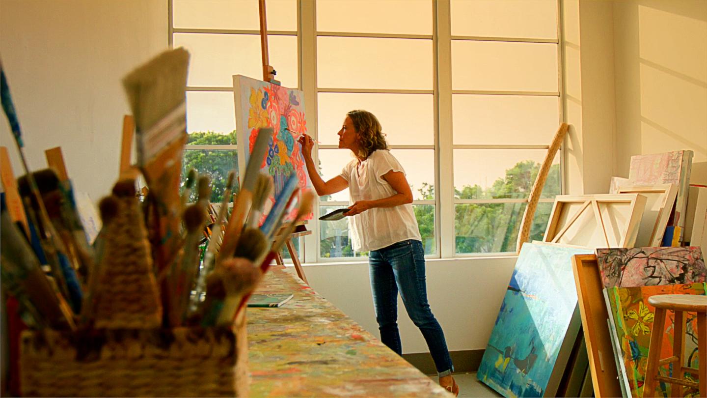 Artist painting a canvas in a sunlit studio with brushes in the foreground.