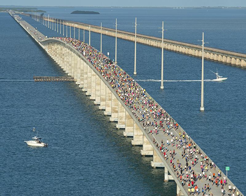 Crowd runs on a long bridge over blue water, boats nearby.