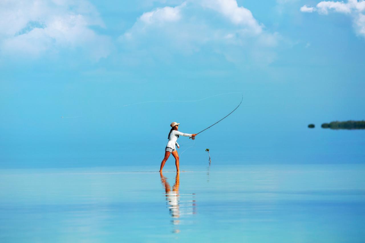 Person fly fishing in calm, shallow blue water under a clear sky.