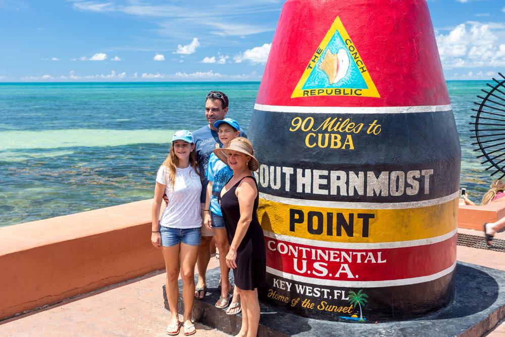 Four people smiling beside the Southernmost Point buoy in Key West, sunny day.