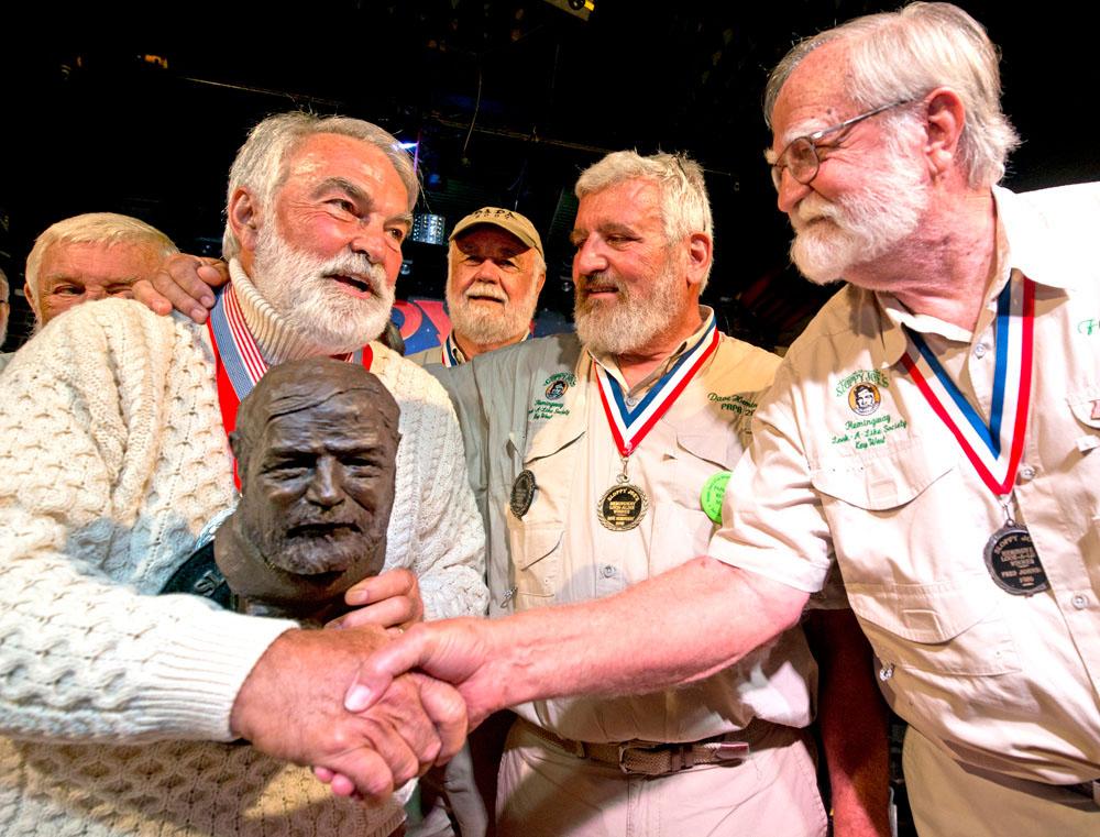 Elderly men with medals gather, one holding a bust, shaking hands, smiling warmly.