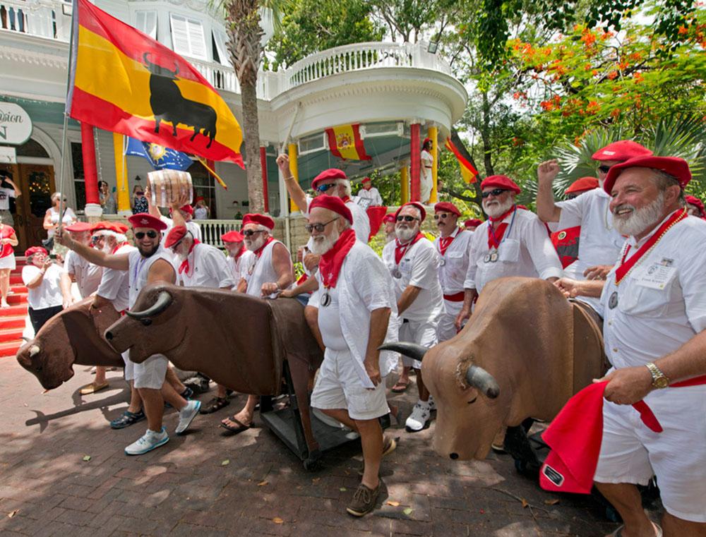 People dressed in white with red sashes and hats, posing with fake bulls, in a festive setting.