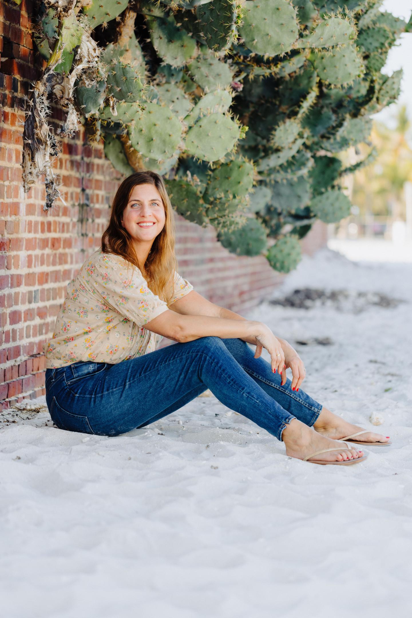 Smiling woman sitting on sandy ground by a brick wall with cactus.