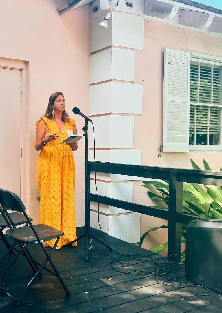 Woman in yellow dress speaks at an outdoor microphone on a sunny terrace.