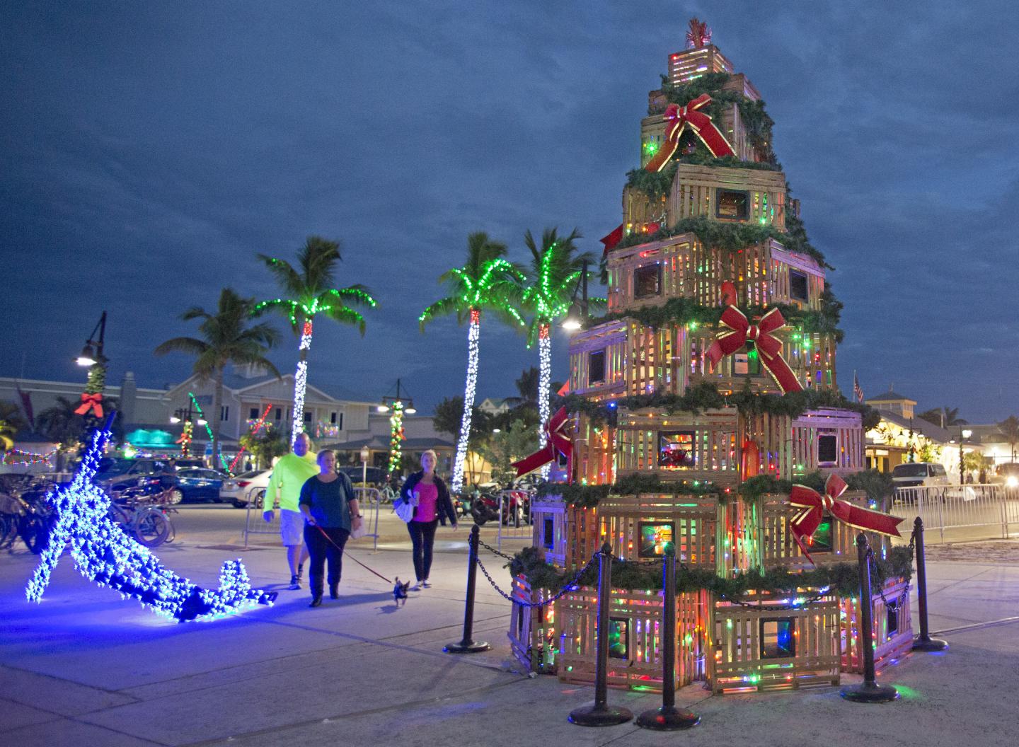 Festive crate Christmas tree with lights, palm trees, and people strolling at dusk.