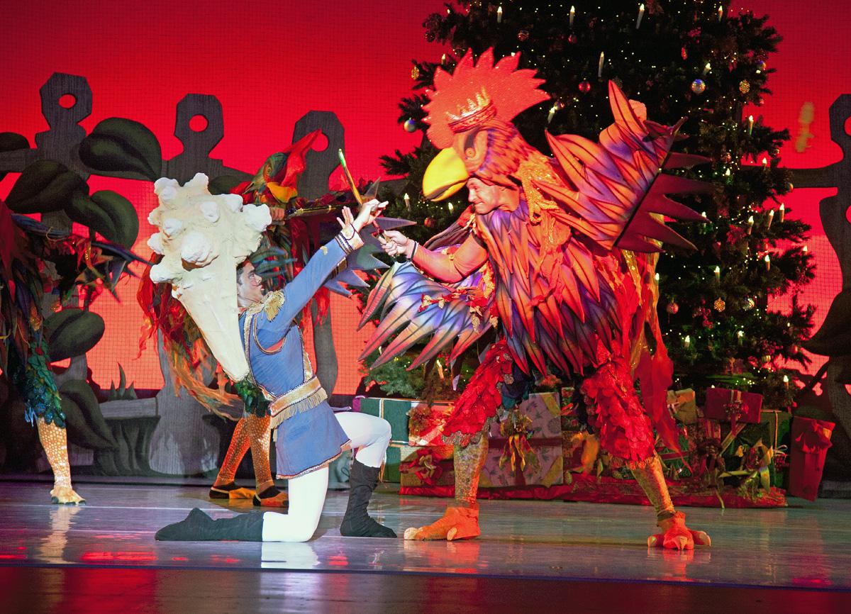 Two performers in colorful costumes dance on stage, with a decorated tree in the background.