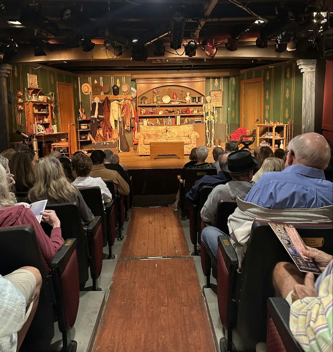 Theater audience facing stage with colorful set and props.
