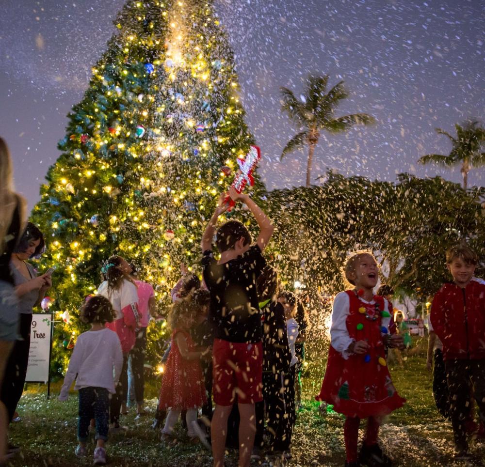 Children excitedly play in snow by a lit Christmas tree at night.