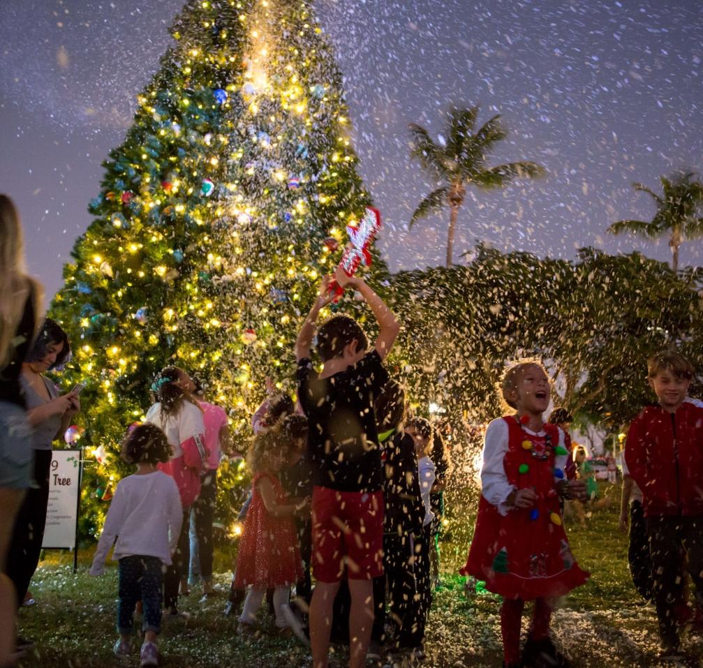 Children playing under artificial snow near a lit Christmas tree at night.