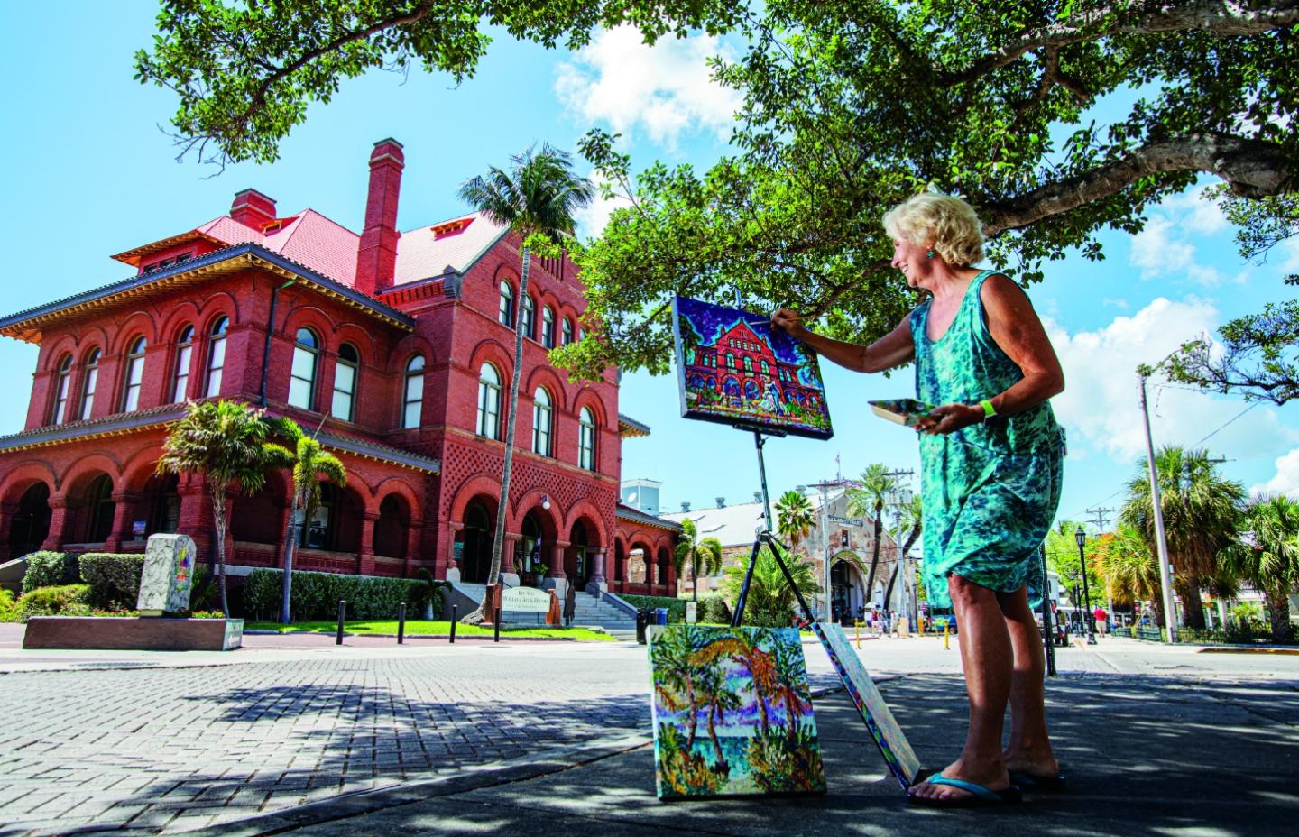 Artist painting a red brick building outdoors under a tree.