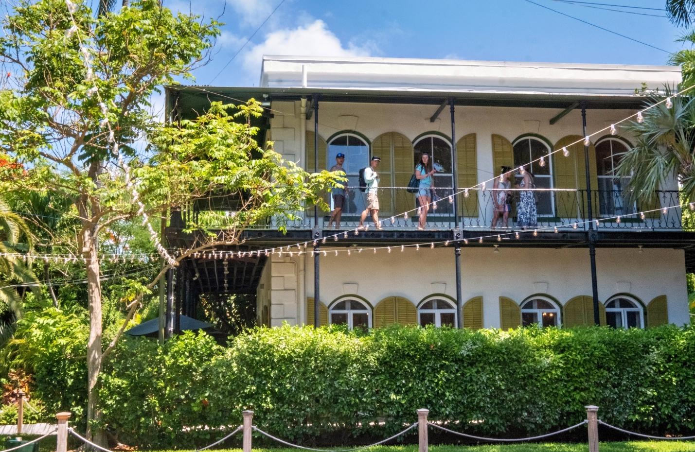 Historic two-story house with a balcony, surrounded by greenery.