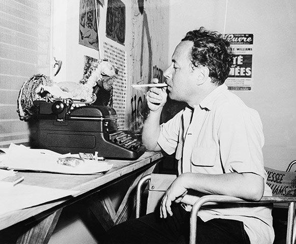 Man seated at a desk typing, smoking a cigarette, black and white photograph.