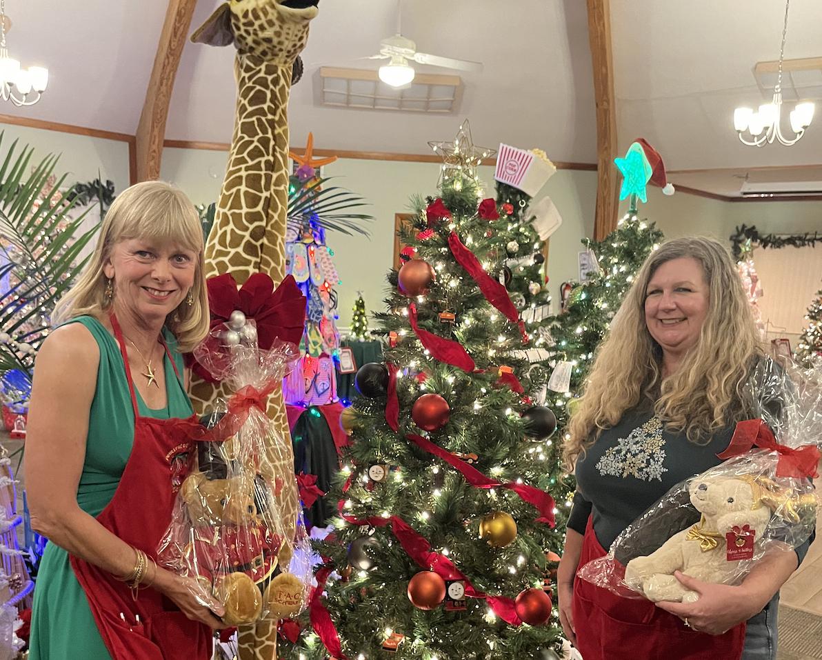 Two women smiling, holding gifts in front of decorated Christmas trees.