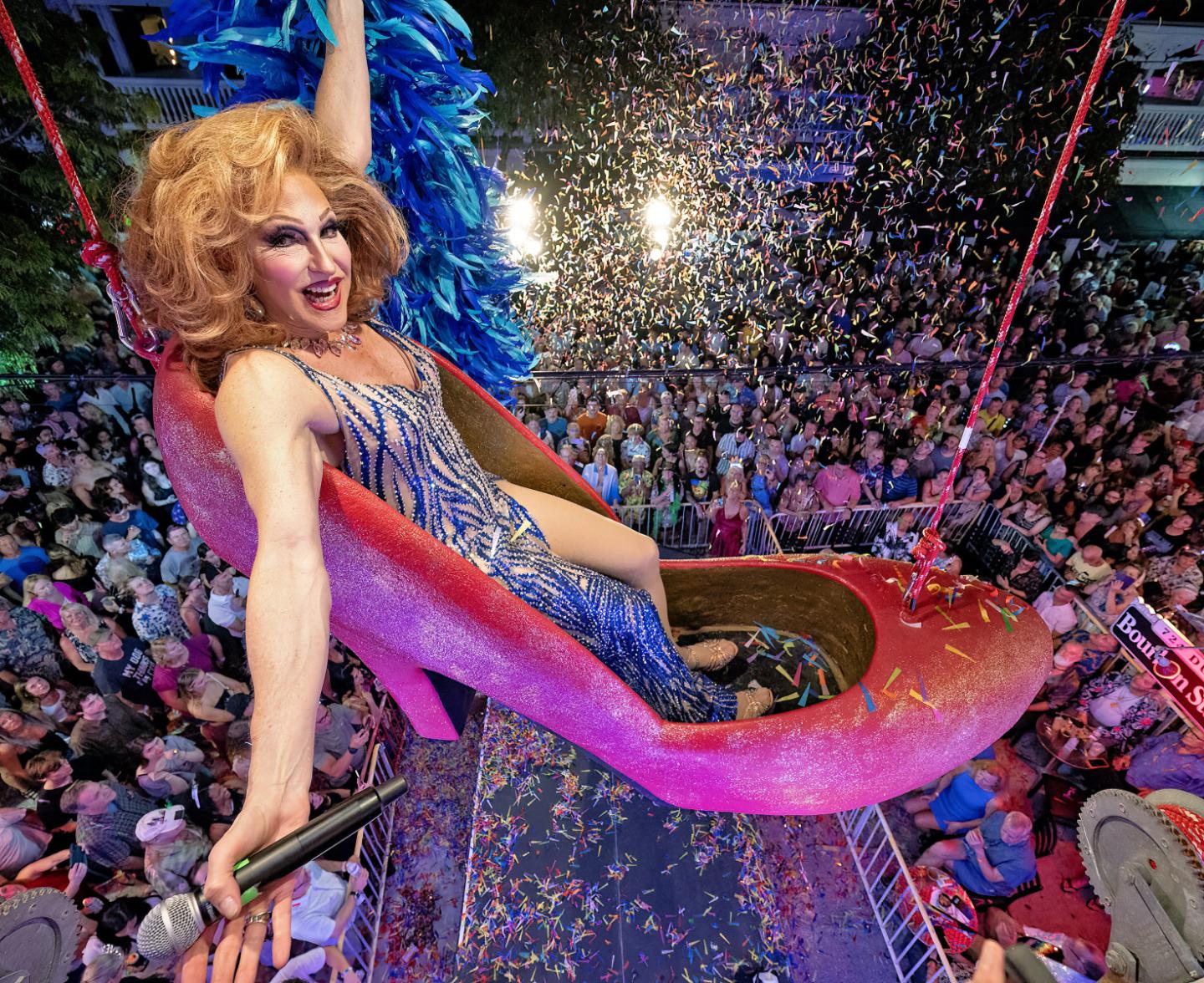 Performer in a giant shoe above a crowd, colorful confetti fills the air.