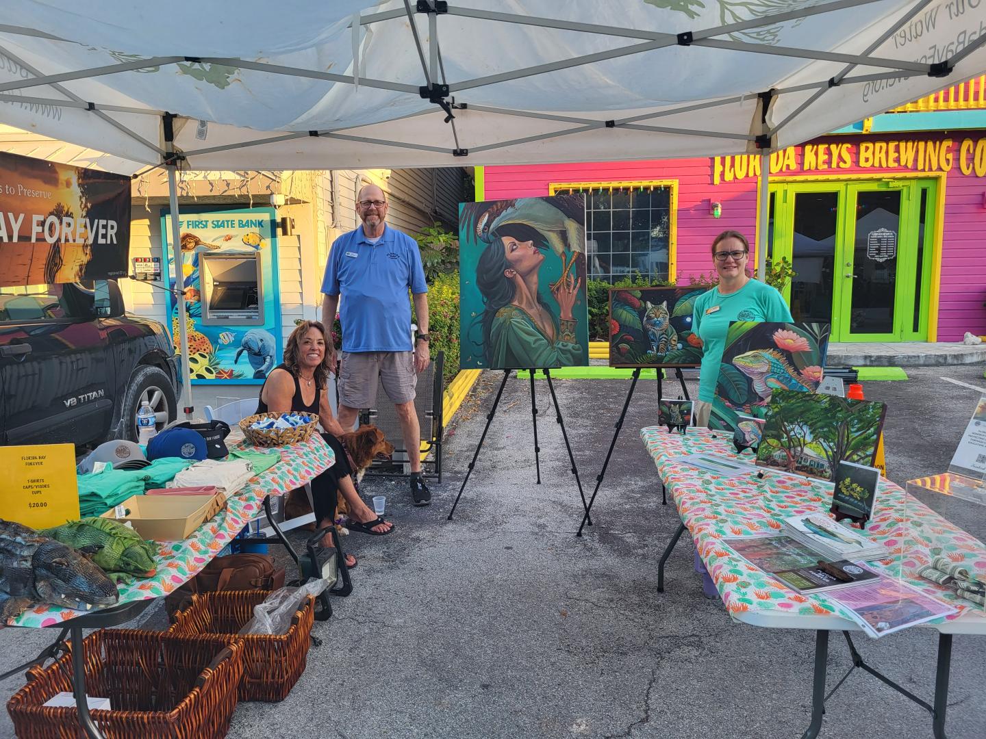 Outdoor art sale with paintings displayed on easels, people seated, vibrant building in background.