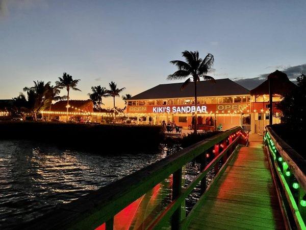 Boardwalk at dusk leading to a lit-up waterfront restaurant with palm trees.