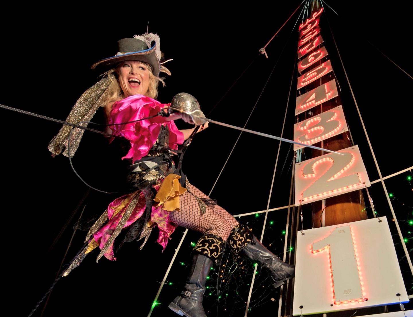 Performer in bright, eclectic outfit on a high wire at night, with a countdown tower behind.