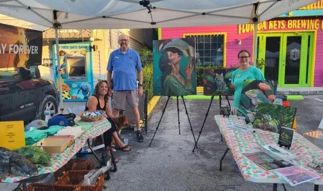 Outdoor art stall with three people and colorful paintings under a tent.