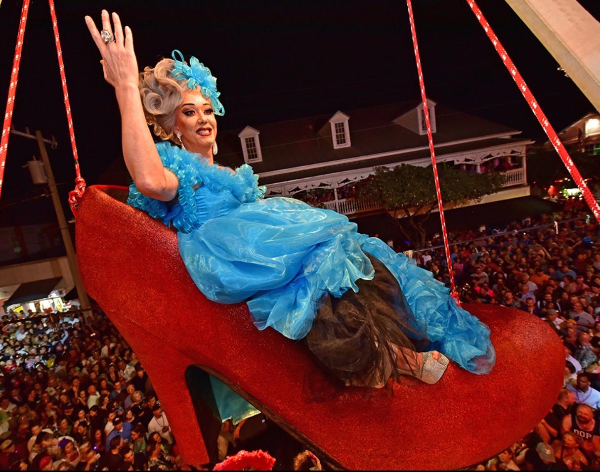 Drag performer in blue gown, smiling and waving, sits in a giant red shoe above a crowd at night.
