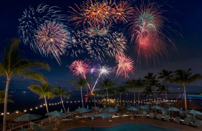 Fireworks explode over a tropical beach at night with palm trees and lights.