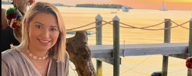Smiling woman at a dock during sunset, sailboats in the distance.
