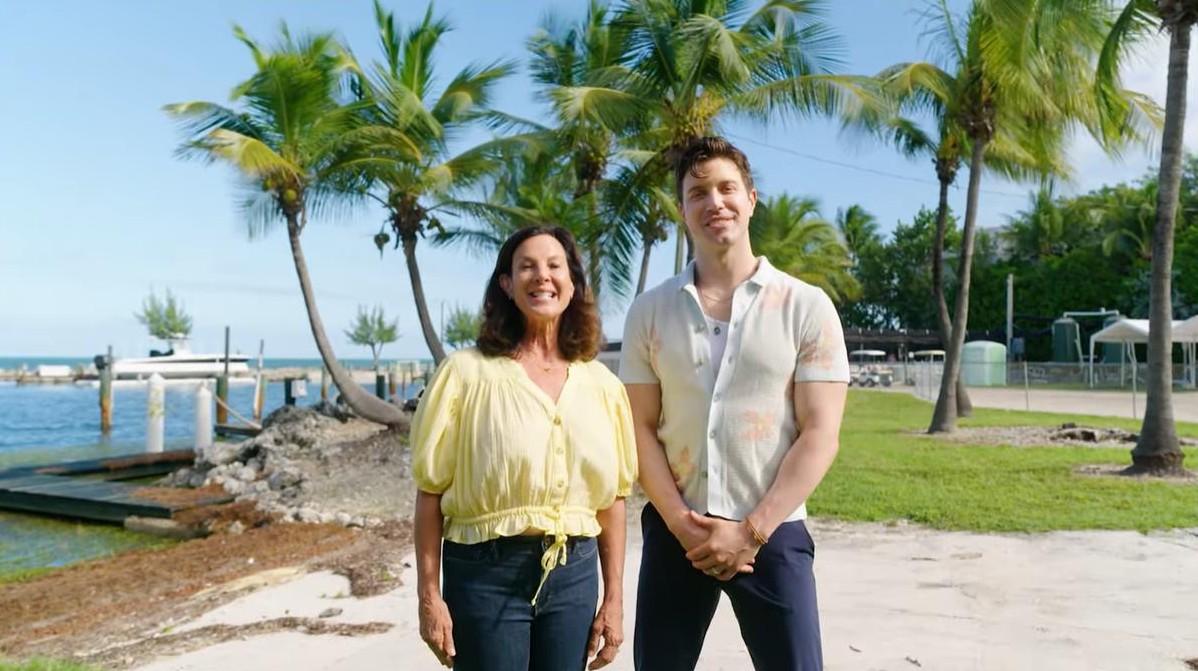 A smiling woman and man stand on a sunny beach with palm trees.