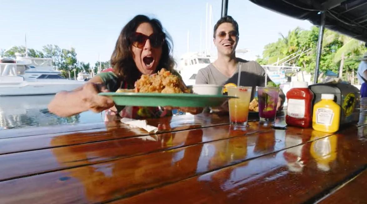 Woman excitedly presenting plate of food at outdoor dining table by the water.