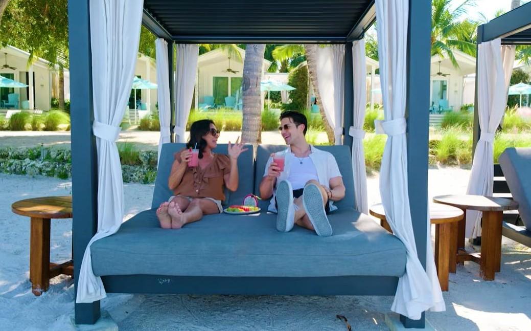 Two people relaxing on a beach cabana, holding drinks and smiling.